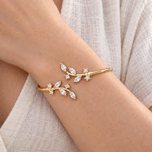 May include: A gold-toned open cuff bracelet featuring two symmetrical branch-like ends. Each branch is adorned with marquise-cut and round-cut clear white stones arranged to resemble leaves and small buds along the polished metal frame.