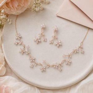 May include: A rose gold necklace and matching drop earrings set featuring delicate vine-like branches adorned with clear marquise-cut crystals arranged in small flower clusters. The jewelry set displays a metallic finish and sparkling stones.