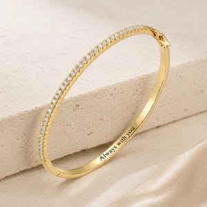 May include: A gold-toned hinged bangle bracelet featuring a row of small, clear, sparkling stones along the outer edge. The inner surface of the metal band is engraved with the black text Always with you, and it includes a secure clasp mechanism.