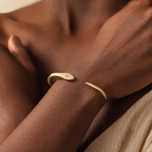 Può includere: Un bracciale aperto color oro a forma di serpente, caratterizzato da una finitura liscia e lucida e da una piccola pietra preziosa rotonda e trasparente incastonata nella testa del serpente. Il bracciale ha un design affusolato verso la coda.