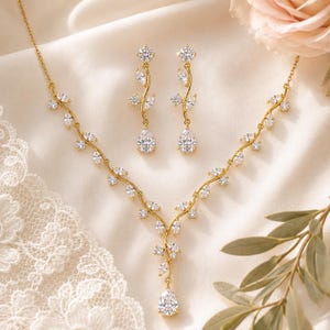 Puede incluir: Un conjunto de collar y pendientes colgantes en tono dorado con un delicado diseño de enredadera. Las piezas están adornadas con piedras transparentes de talla marquesa en forma de hoja, con una piedra central en forma de pera.