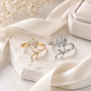 Può includere: Due anelli aperti con design floreale e pietre trasparenti. Un anello è color oro con motivo a fiore e foglie, mentre l'altro è color argento con un design a fiore e foglie abbinato, entrambi decorati con scintillanti cristalli trasparenti.
