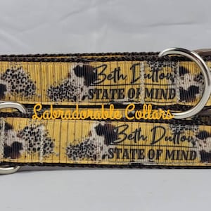 May include: A yellow and brown dog collar with a black and white print of a hedgehog. The collar has a brown buckle and a silver D-ring. The text "Beth Dutton State of Mind" is printed on the collar.