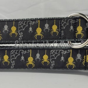 Może przedstawiać: Dwa czarne i szare obroże dla psa z wzorem pajęczyny i pająka. Obroże mają srebrne metalowe kółka D i szare plastikowe klamry.