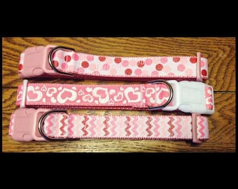 May include: Three pink dog collars with different patterns. The collars have pink and white hearts, pink and white polka dots, and pink and white zigzags.