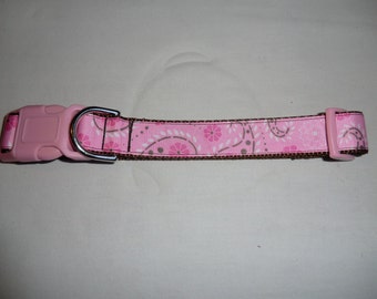 May include: Pink dog collar with a brown and white floral pattern. The collar has a pink plastic buckle and a silver metal D-ring.