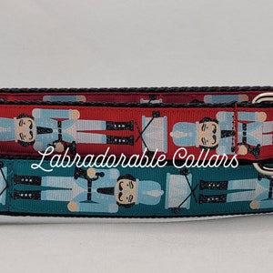 May include: Two dog collars with a nutcracker pattern. The top collar is red with white accents and the bottom collar is teal with white accents. Both collars have black buckles and silver D-rings. The text "Labradorable Collars" is printed on the collars.