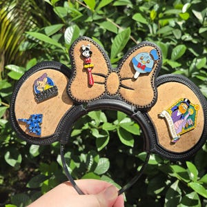 May include: Black headband with cork ears and a cork bow. The ears and bow are decorated with colorful character pins. The pins include Mickey Mouse, Donald Duck, and other Disney characters. The headband is held by a hand in front of a green leafy background.