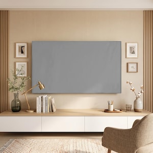 Op de afbeelding: Een moderne woonkamer met een groot grijs scherm aan een lichtgekleurde muur. Onder het scherm staat een houten console met boeken, een lamp en decoratieve items. Een beige fauteuil en een patroonkleed maken de scène compleet.