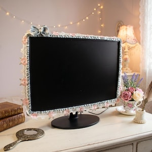 Peut inclure: Un écran d'ordinateur avec une housse décorative. La housse est bleu clair avec une bordure en dentelle et des roses roses. Un nœud bleu clair est placé en haut. L'écran est posé sur un bureau blanc avec un miroir et des livres vintage.