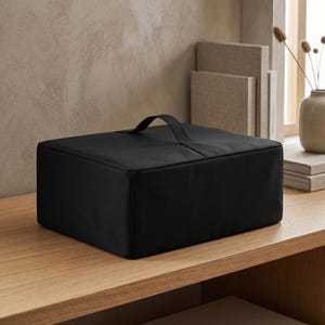 Puede incluir: Una caja de almacenamiento negra con un asa en la parte superior. La caja rectangular está colocada sobre una superficie de madera. El fondo incluye una ventana, libros y un jarrón con plantas secas.