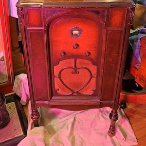 May include: A vintage floor-standing wooden radio cabinet featuring a dark brown finish, a central speaker grille with a heart-shaped cutout, three circular control knobs, and a small illuminated dial display at the top, supported by four carved legs.