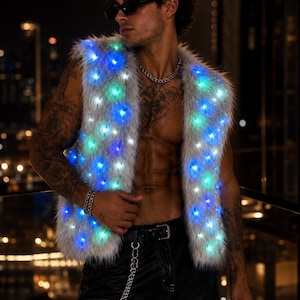 Puede incluir: Un chaleco sin mangas de piel sintética gris claro con luces LED integradas que brillan en tonos azules y verdes. El chaleco se lleva abierto, mostrando un material texturizado y peludo con un pelo denso y puntos de luz espaciados uniformemente.