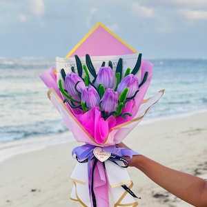 Puede incluir: Un ramo de tulipanes morados envuelto en papel rosa y blanco, atado con una cinta morada y una etiqueta en forma de corazón que dice "Valentine's Day". El fondo es una playa con el océano y el cielo.