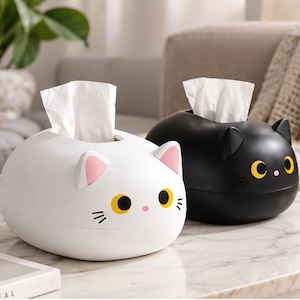 Pode incluir: Duas caixas de lenços ovais com formato de gato. Uma é branca com o interior das orelhas rosa e olhos amarelos, enquanto a outra é preta com olhos amarelos. Ambas possuem bigodes e um pequeno nariz rosa, com lenços brancos saindo do topo.