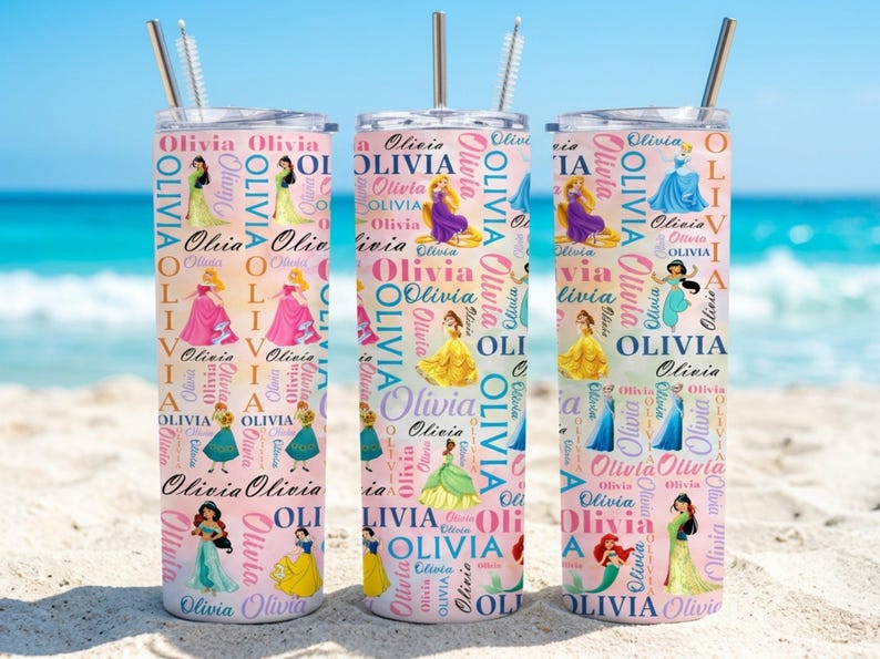 May include: Three personalized tumblers with princess illustrations and the name "Olivia" repeated in a pink and white pattern. Each tumbler has a silver straw and a lid. The background shows a beach and ocean.