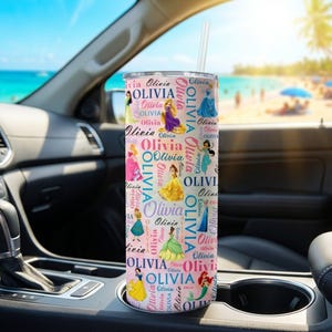 May include: A pink tumbler with a straw, featuring the name "Olivia" in a repeating pattern with princess illustrations. The tumbler is cylindrical and appears to be stainless steel. The background shows a car interior and a beach scene.