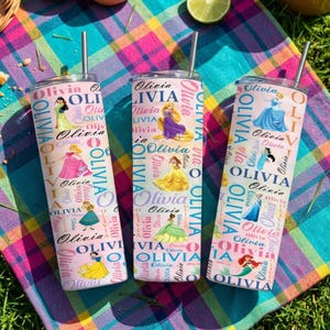 May include: Three stainless steel tumblers with straws, featuring princess illustrations and the name "Olivia" in a repeating pattern. The tumblers are on a colorful plaid blanket, with a lime and other items in the background.