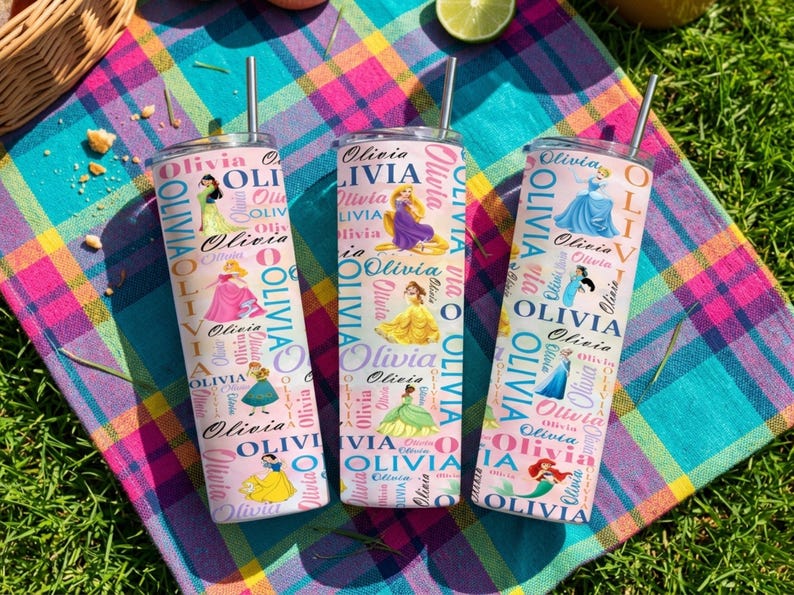 May include: Three stainless steel tumblers with straws, featuring princess illustrations and the name "Olivia" in a repeating pattern. The tumblers are on a colorful plaid blanket, with a lime and other items in the background.