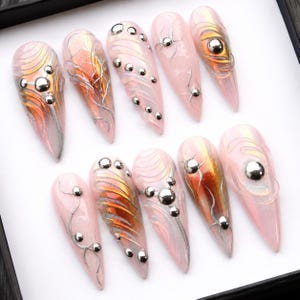 May include: A set of ten stiletto-shaped press-on nails featuring a translucent pale pink base with iridescent orange and gold swirling accents. Each nail is decorated with raised silver metallic lines and small polished silver spherical studs.