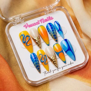 May include: A set of ten artificial nails in a clear plastic case. The nails feature various designs, including yellow with blue swirls, orange and blue gradients, and gold accents. The text "Veucci Nails" and "Thank you for your order" are visible.
