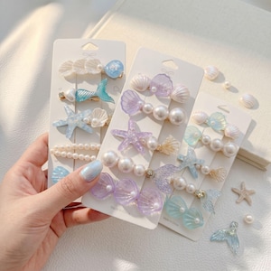 May include: Several hair clip sets featuring mermaid and ocean-themed designs. Each set includes clips shaped like seashells, starfish, and mermaid tails, in shades of blue, purple, and white. Some clips have pearl accents.