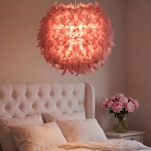 May include: A pink feather pendant light hangs above a bed with white linens and a pink throw. The light fixture is round and fluffy, casting a warm glow. A bouquet of pink flowers sits on a bedside table.