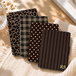 May include: Four rectangular e-reader cases arranged in a row. The first features a brown and black leopard print, the second a black and beige plaid pattern, the third a brown cover with white polka dots, and the fourth a brown striped case with the gold initials RS.