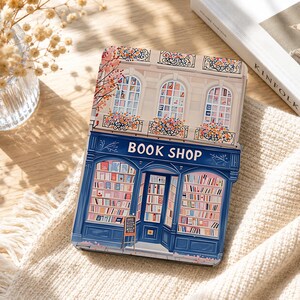 Könnte beinhalten: Eine rechteckige E-Reader-Hülle mit einer aufgedruckten Illustration einer Buchhandlungsfassade. Das Design zeigt ein dunkelblaues Ladenlokal mit großen, mit Bücherregalen gefüllten Fenstern, einer zentralen Tür und blumengeschmückten Fenstern.