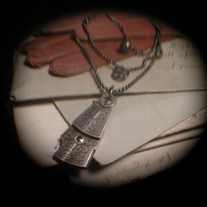 Puede incluir: Un collar de plata con un colgante de diseño detallado y ornamentado. El colgante tiene la forma de una pequeña casa triangular con una pequeña piedra transparente en el centro.