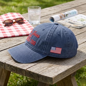 May include: A blue baseball cap with the text "Jesus and America Too" embroidered in red. The cap features a small American flag patch and a curved brim. The hat is sitting on a wooden picnic table.