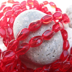 May include: A strand of red glass beads, oval shaped, with a faceted finish. The beads are arranged in a loose pile on a gray, textured surface.
