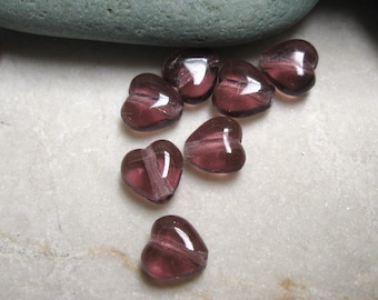 May include: A collection of seven heart-shaped beads in a deep, translucent purple hue. The beads are scattered on a light-colored surface, with a smooth, glossy finish. The beads are approximately 0.5 inches in size.