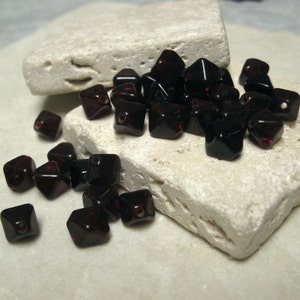 May include: A collection of dark red, square-shaped beads with faceted edges. The beads are scattered on a light-colored, textured surface. Each bead has a small hole, suggesting they are for stringing. The beads have a glossy finish.