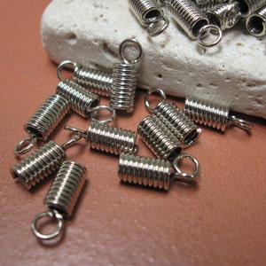 May include: Close-up of several silver-colored spring clasps with small loops at the ends. The clasps are cylindrical and coiled, designed for jewelry making or crafts. They are scattered on a light-colored, textured surface.