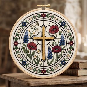 Pode incluir: Bordado circular com uma cruz dourada e motivos florais. O desenho inclui papoilas vermelhas, tremoços azuis e outras flores, tudo dentro de um bastidor de bordado de madeira. O tecido é de cor creme.