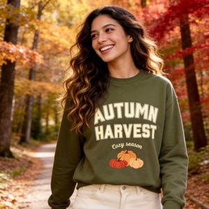 Puede incluir: Una sudadera de cuello redondo color verde oliva que presenta el texto en color crema "AUTUMN HARVEST" en una fuente arqueada en negrita, seguido del texto en cursiva "cozy season" y una ilustración gráfica de tres calabazas en tonos naranja, rojo y crema.