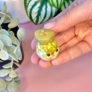 Pode incluir: Pequena e fantasiosa escultura em cerâmica de uma criatura redonda sorrindo com um picles na cabeça. A escultura é principalmente amarela e verde com olhos pretos e boca rosa. O fundo é rosa claro.
