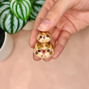 以下が含まれることがあります： 茶色の斑点、濃い赤色の目、開いたピンク色の口を持つ、丸みを帯びたベージュ色の生き物が2つ重なった小さな陶器の置物。上の生き物は下の生き物よりも小さく、どちらも滑らかで光沢のある仕上がりになっています。