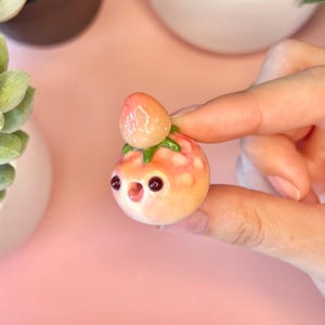 May include: A small, round, peach-colored sculpture of a strawberry with a face, held between fingers. The strawberry has a green stem and a smaller strawberry on top. The sculpture has two dark eyes and a small mouth.