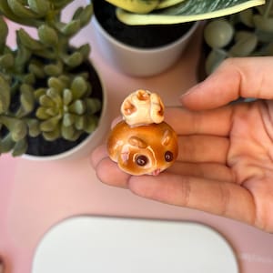 Könnte beinhalten: Eine kleine, glänzende, hellbraune und braune Keramikfigur in Form eines Hamsters mit einem winzigen, hellen, strukturierten Objekt auf dem Kopf. Die Figur hat dunkelbraune Augen, kleine Ohren und einen glatten, abgerundeten Körper mit glänzendem Finish.