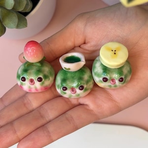 May include: Three small, round, green-and-white ceramic figurines with expressive faces, each topped with a different miniature object: a pink peach, a tiny white bowl filled with green liquid, and a yellow banana slice.