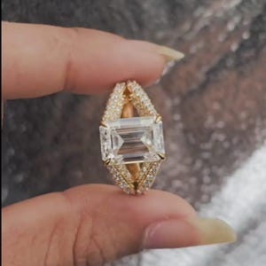 Pode incluir: Um anel de ouro com uma grande pedra preciosa retangular e transparente no centro. O anel é rodeado por pedras preciosas menores e transparentes e tem um design de haste dividida. O anel é segurado entre os dedos.
