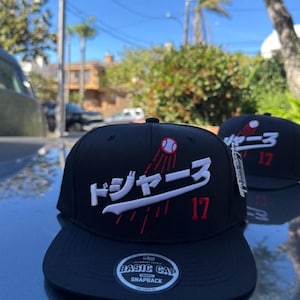 May include: A black snapback cap featuring white Japanese text and a white baseball bat graphic across the front. A red and white baseball icon is positioned above the text, with the red number 17 embroidered below. A circular sticker is on the brim.