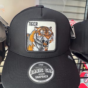 May include: A black snapback cap featuring a rectangular patch with an embroidered tiger graphic and the word TIGER above it. The cap has a black mesh back and a circular sticker on the brim that reads BASIC CAP ORIGINAL SNAPBACK LOS ANGELES.
