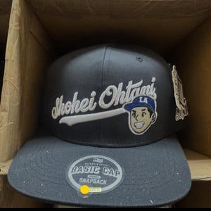 May include: A dark gray snapback cap featuring the embroidered white text Shohei Ohtani and a cartoon face graphic wearing a blue LA cap. A circular sticker on the flat brim reads Basic Cap Original Snapback with a small flag icon and text.