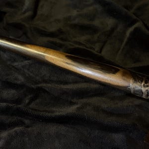 May include: A handcrafted wooden wand with a dark finish and intricate carvings near the handle. The wand is displayed on a wooden stand against a dark background, showcasing its detailed craftsmanship.