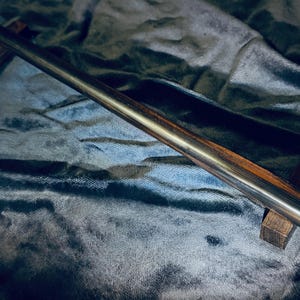 May include: A dark, slender wand with a pointed tip, resting on two small, dark wooden stands. The wand has a polished, metallic appearance with visible wood grain. The background is a dark, textured fabric.