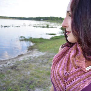 May include: A woman with long dark hair wearing a pink and brown knitted shawl with a beaded trim. The shawl has a lacy pattern and is draped around her shoulders. The woman is looking off to the side and has a thoughtful expression on her face.