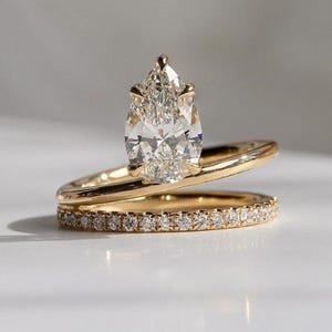 May include: A gold engagement ring with a pear-shaped diamond center stone. The ring is set on a gold band, with a second band below it, encrusted with small diamonds. The rings are displayed on a white surface.